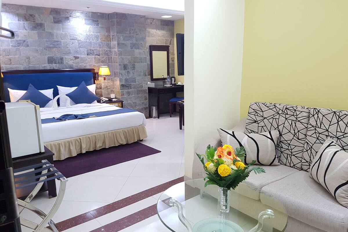 A hotel room with a stone wall, a bed with blue bedding, a desk with a lamp, a small table with a vase of flowers, and a sofa with patterned cushions.