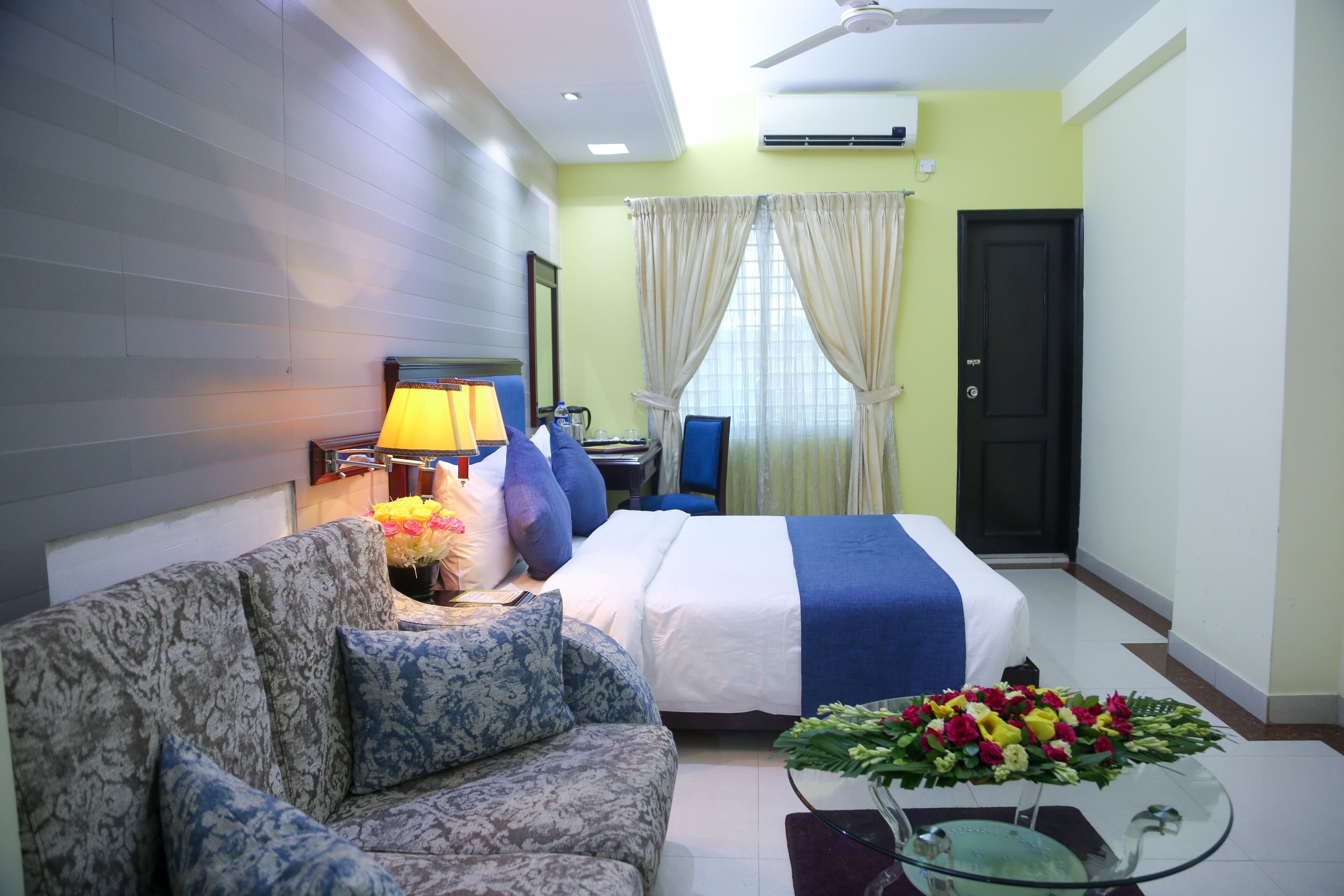 Habitación de hotel con cama, sofá, una mesa de centro de vidrio con flores, lámpara y televisor.