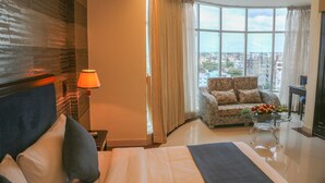 A hotel room with a bed, a sofa, a glass coffee table with flowers, and a view of the city through large windows. - Hotel Valley Garden (Sylhet)