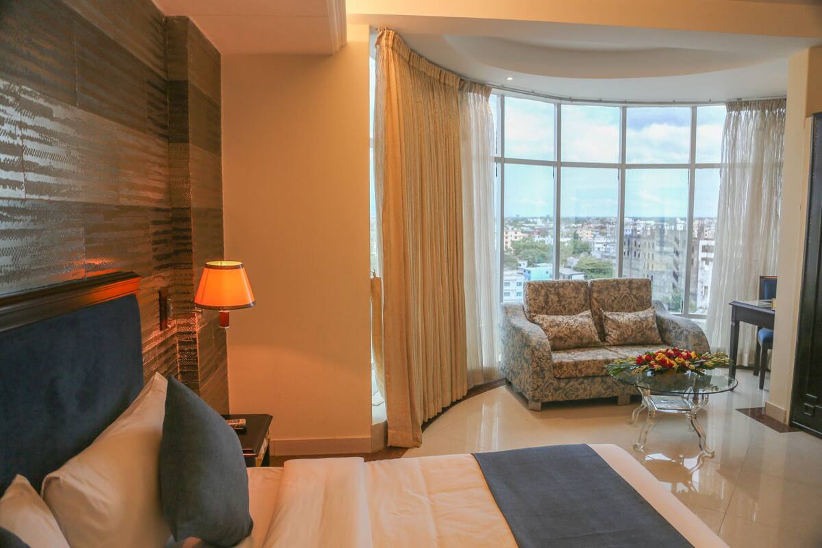 A hotel room with a bed, a sofa, a glass coffee table with flowers, and a view of the city through large windows.
