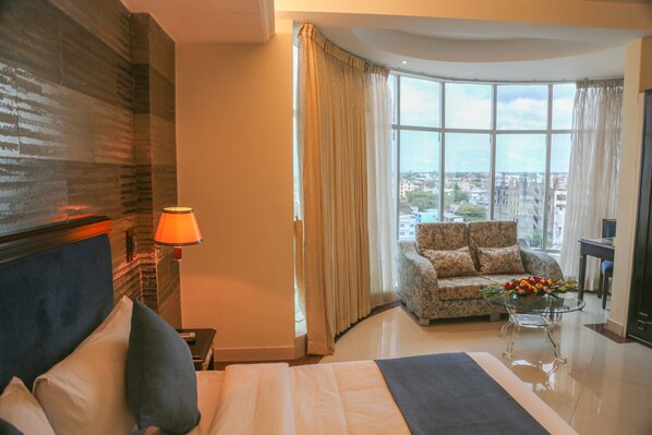 A hotel room with a bed, a sofa, a glass coffee table with flowers, and a view of the city through large windows. - Hotel Valley Garden (Sylhet)