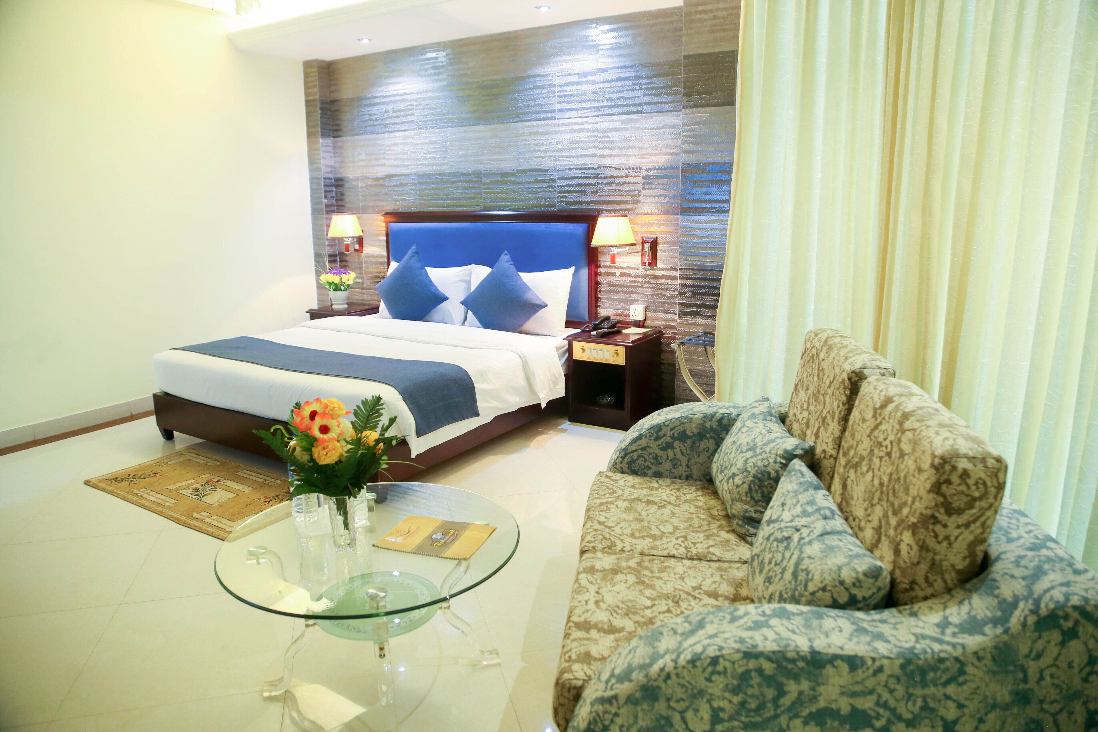 Una habitación de hotel con una cama, una mesita de centro de vidrio con un florero, un sofá estampado y una mesita de noche con una lámpara.