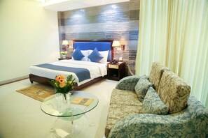 A hotel room with a bed, a glass coffee table with a vase of flowers, a patterned sofa, and a nightstand with a lamp. - Hotel Valley Garden (Sylhet)