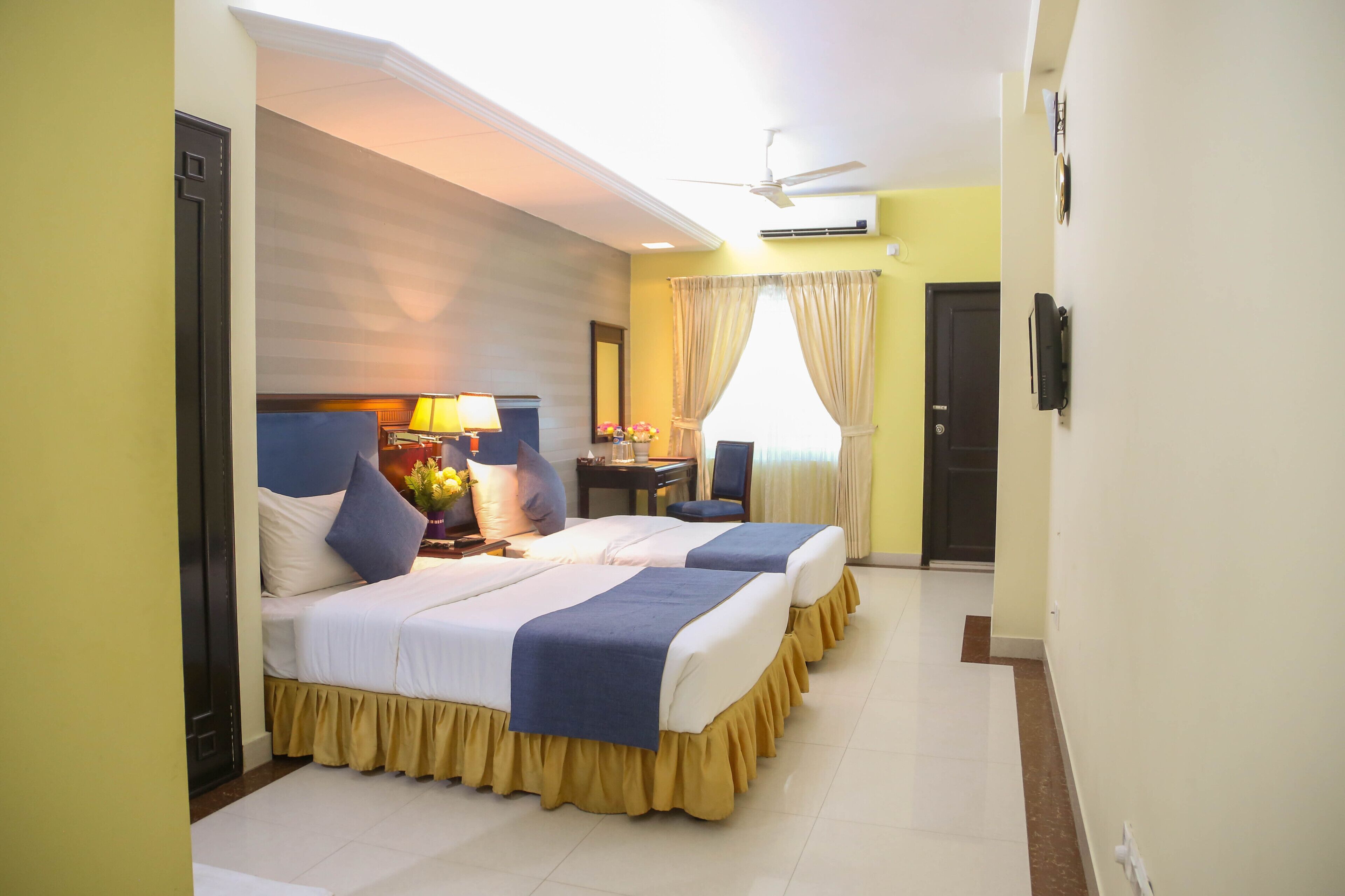 A hotel room with two beds, a desk, a chair, a lamp, and a ceiling fan.