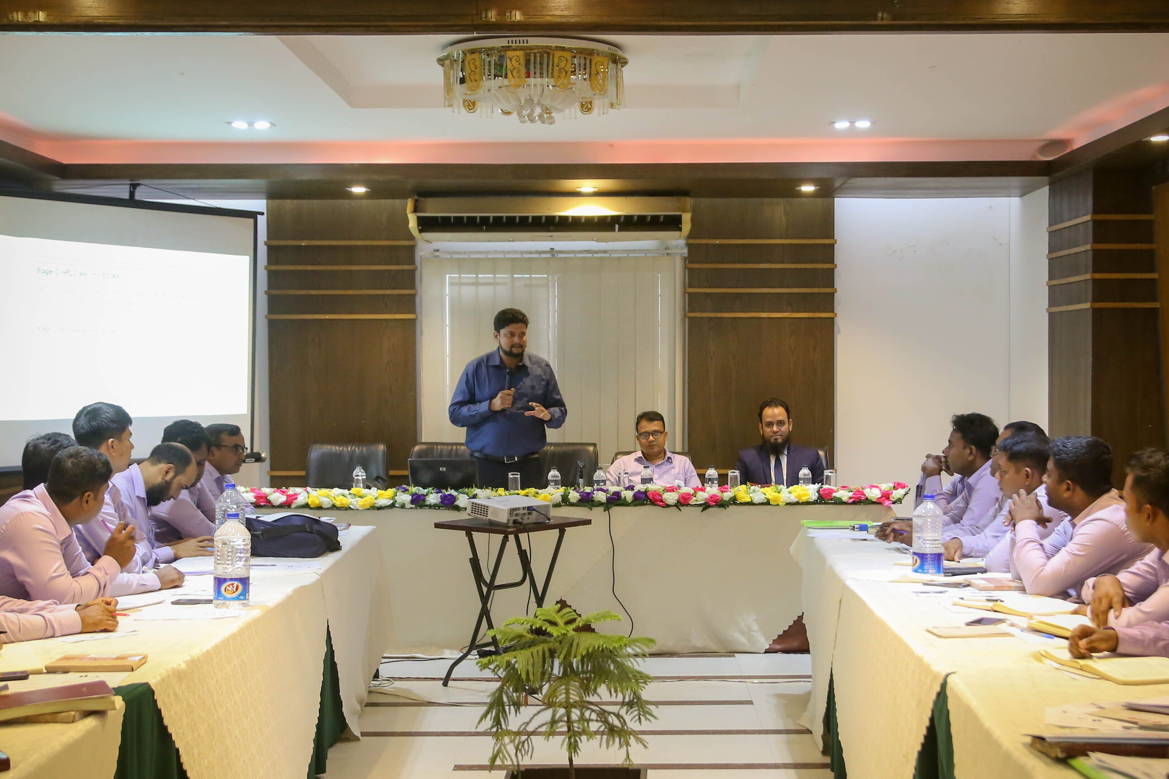 A conference room with a long table, attendees, and a speaker at the front.