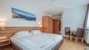 Une chambre d’hôtel moderne avec un grand lit, une tête de lit en bois, un bureau avec deux chaises et un tableau représentant un paysage de montagne accroché au mur.