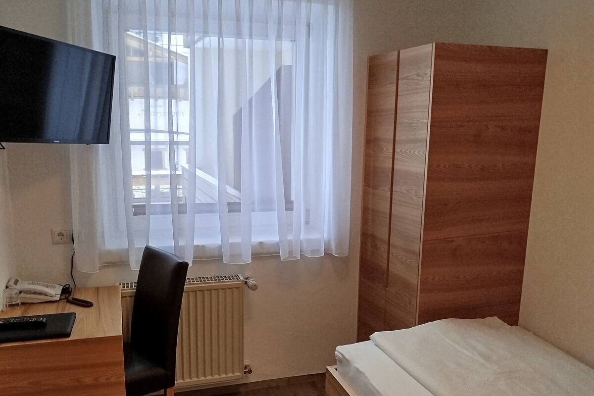A small, well-lit room with a wooden desk, a chair, a bed, and a TV mounted on the wall.