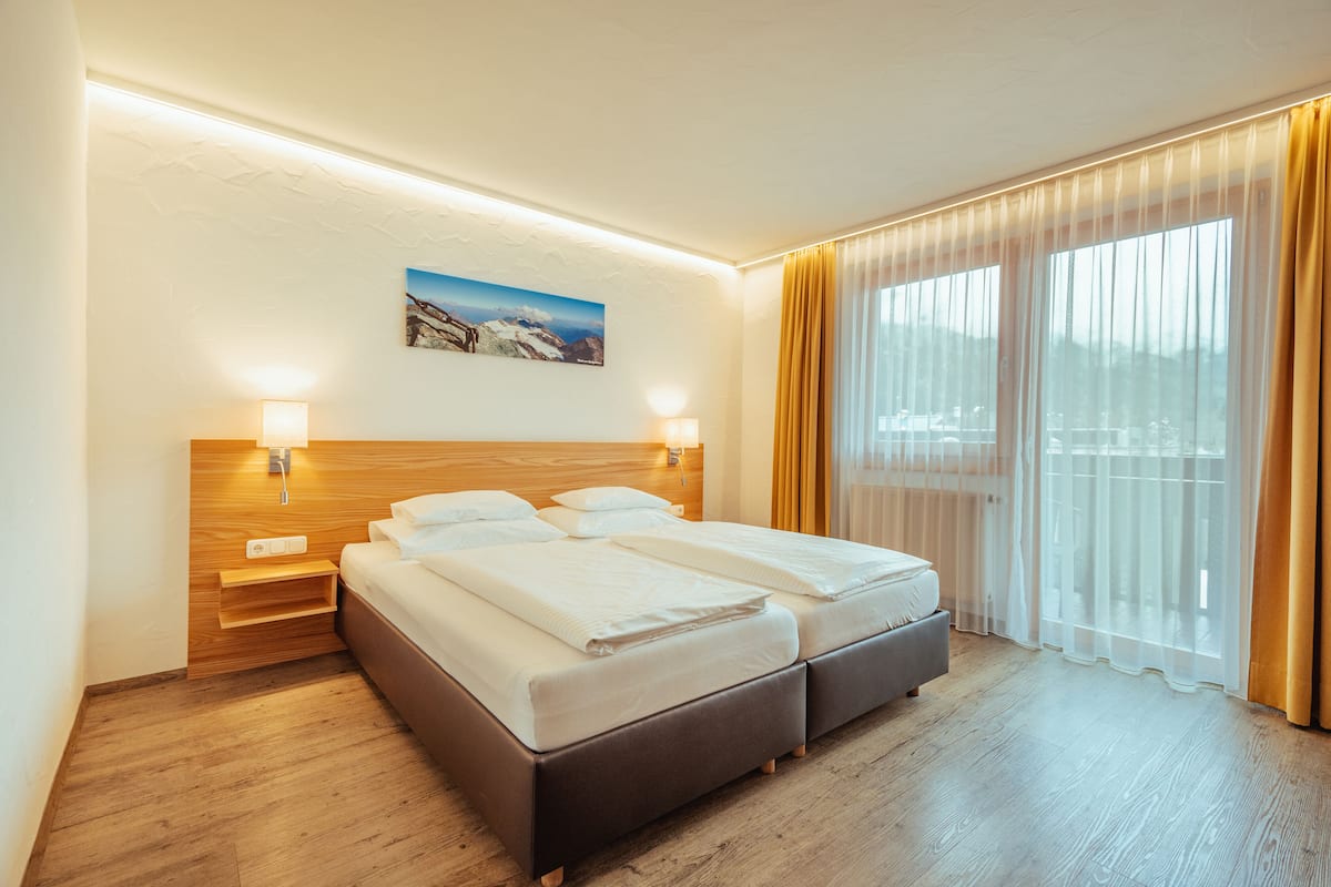 A double bed with a wooden headboard, bedside tables with lamps, a painting on the wall, and a large window with curtains.