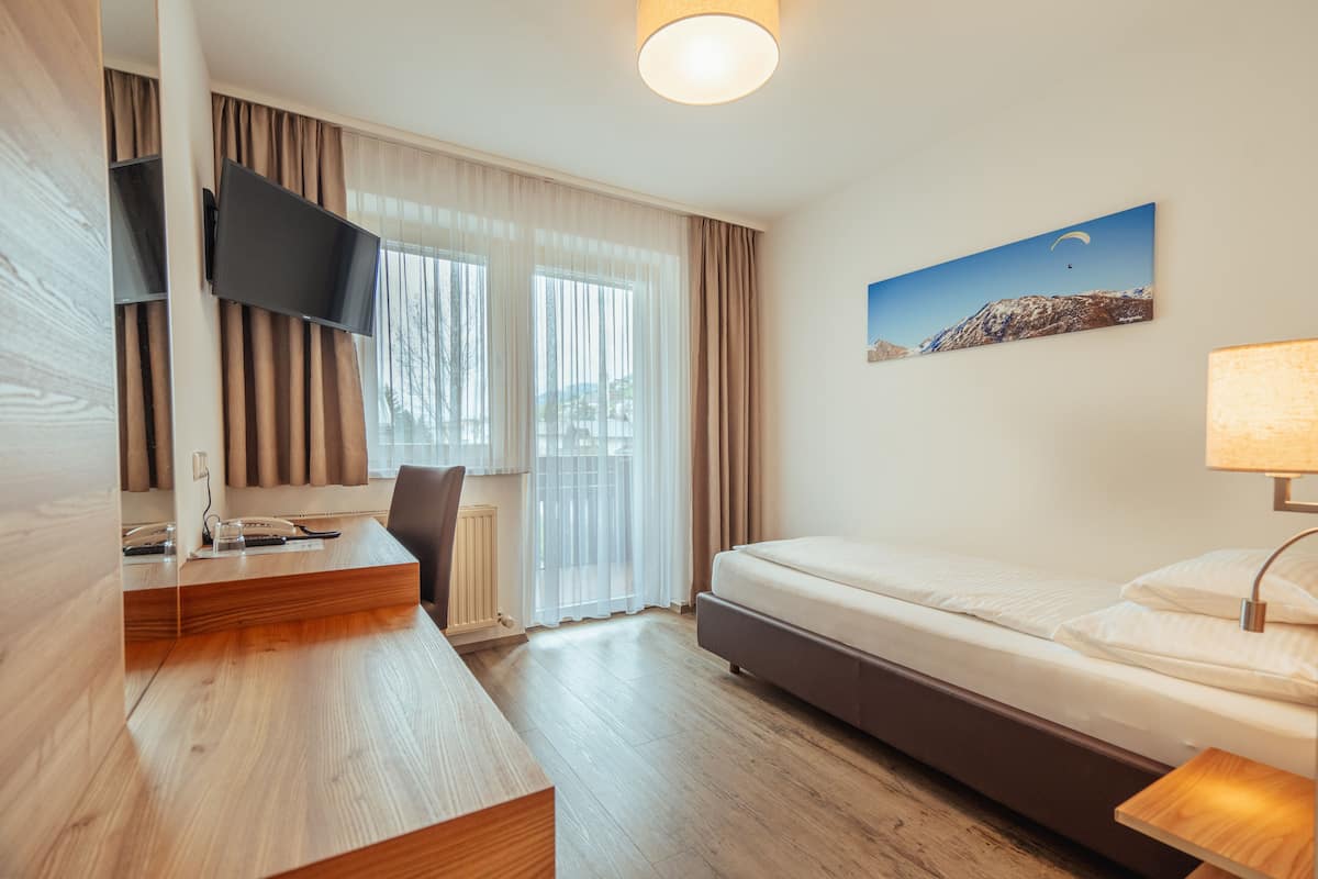 A modern hotel room with a wooden desk, a flat-screen TV, a bed, and a painting of a mountain landscape.