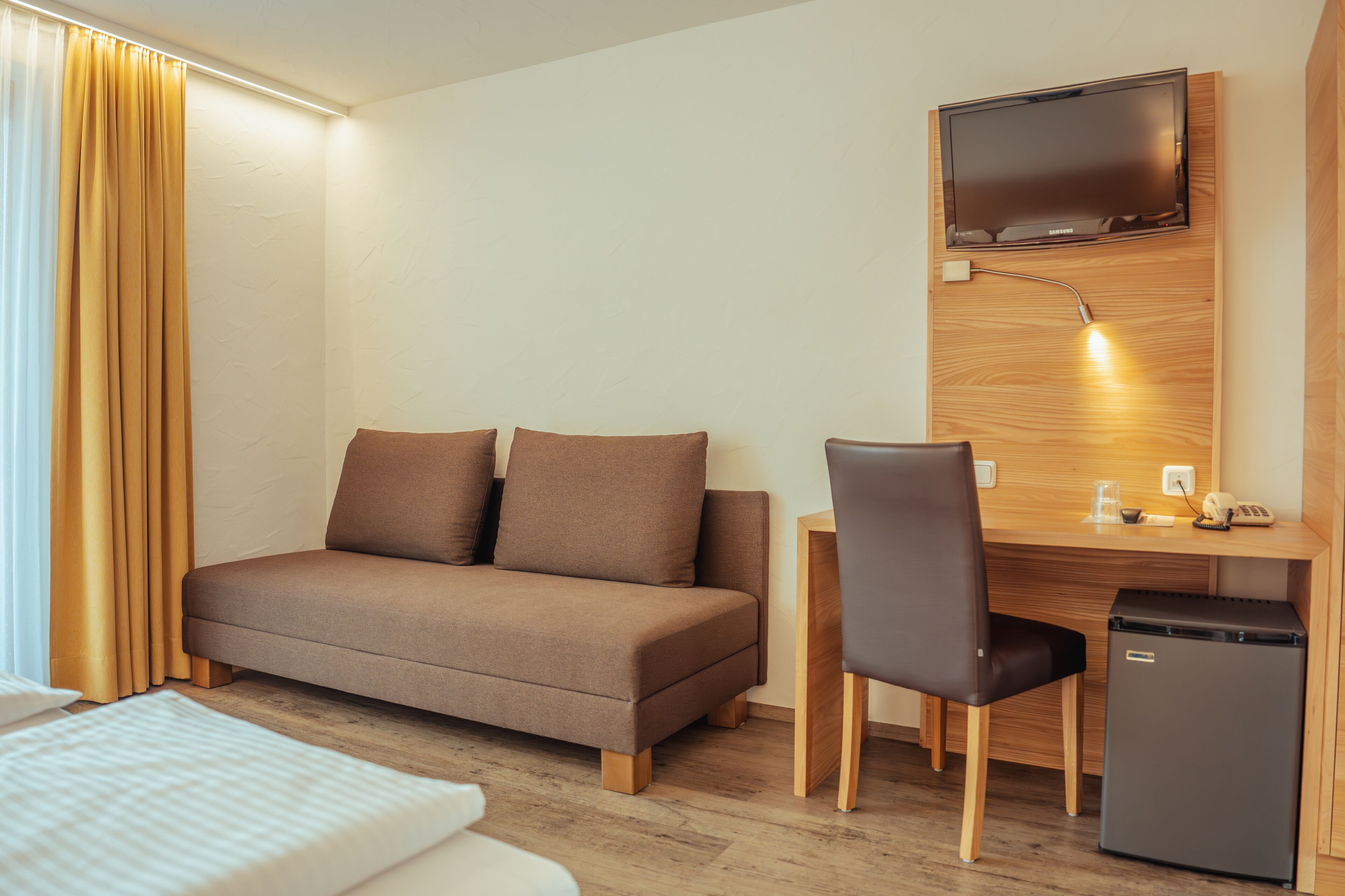 A hotel room with a sofa, a desk with a chair, a television mounted on the wall, and a small refrigerator.