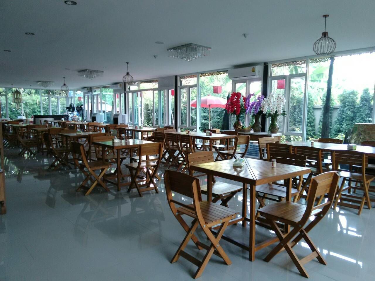 A spacious dining area with wooden tables and chairs, large windows with a view of greenery, and pendant lighting.