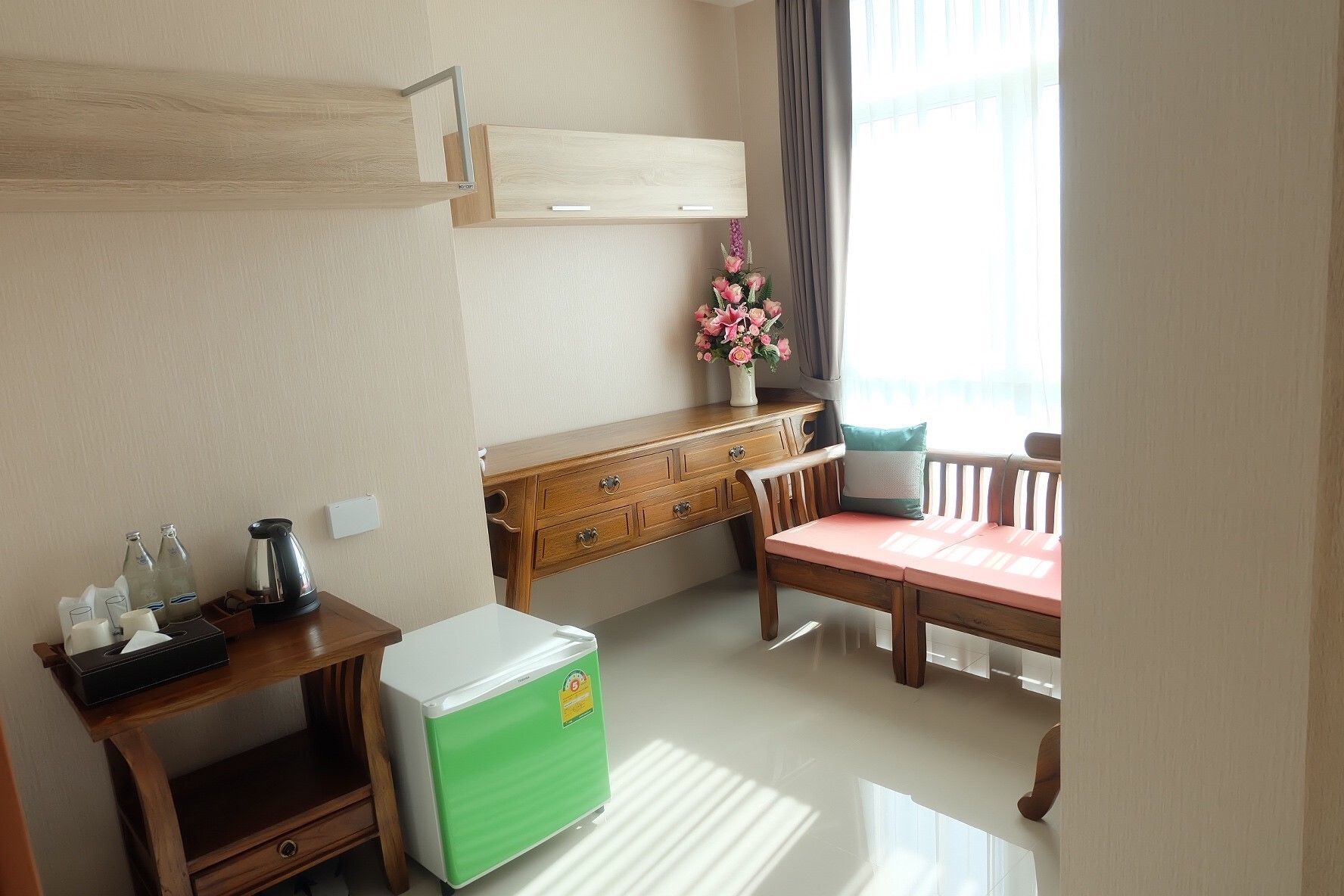 Una habitación compacta con escritorio de madera, refrigerador verde, mesa de comedor con sillas, ventana con cortinas y un jarrón con flores.