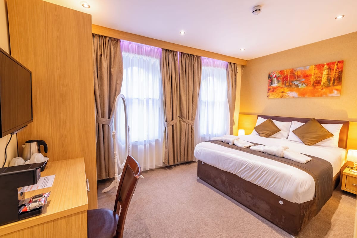 Quarto de hotel com cama, escrivaninha, cadeira, televisão e um quadro na parede.