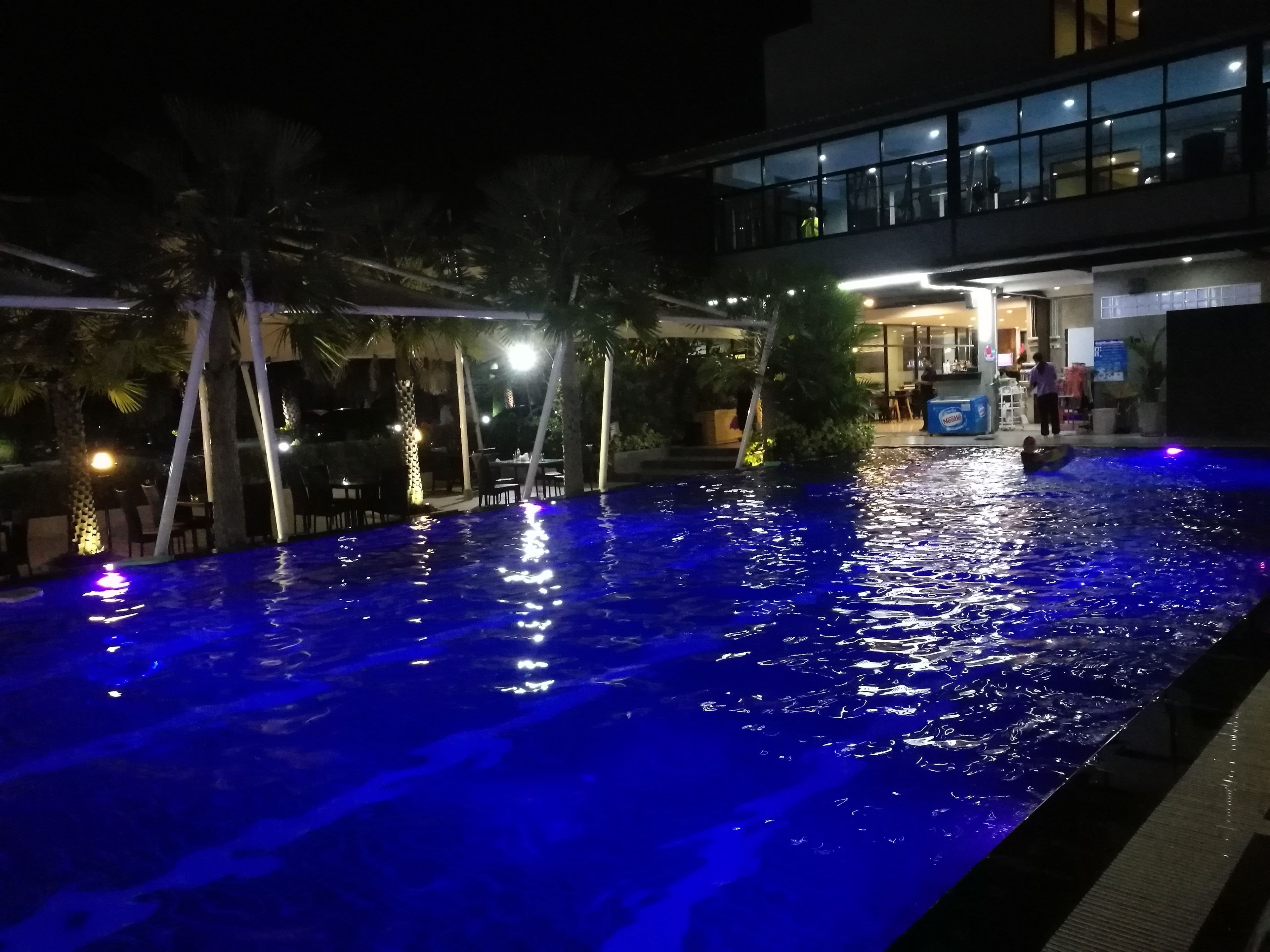 Une vue nocturne d’une piscine illuminée de lumière bleue, entourée de palmiers et d’un bâtiment doté d’une façade en verre.