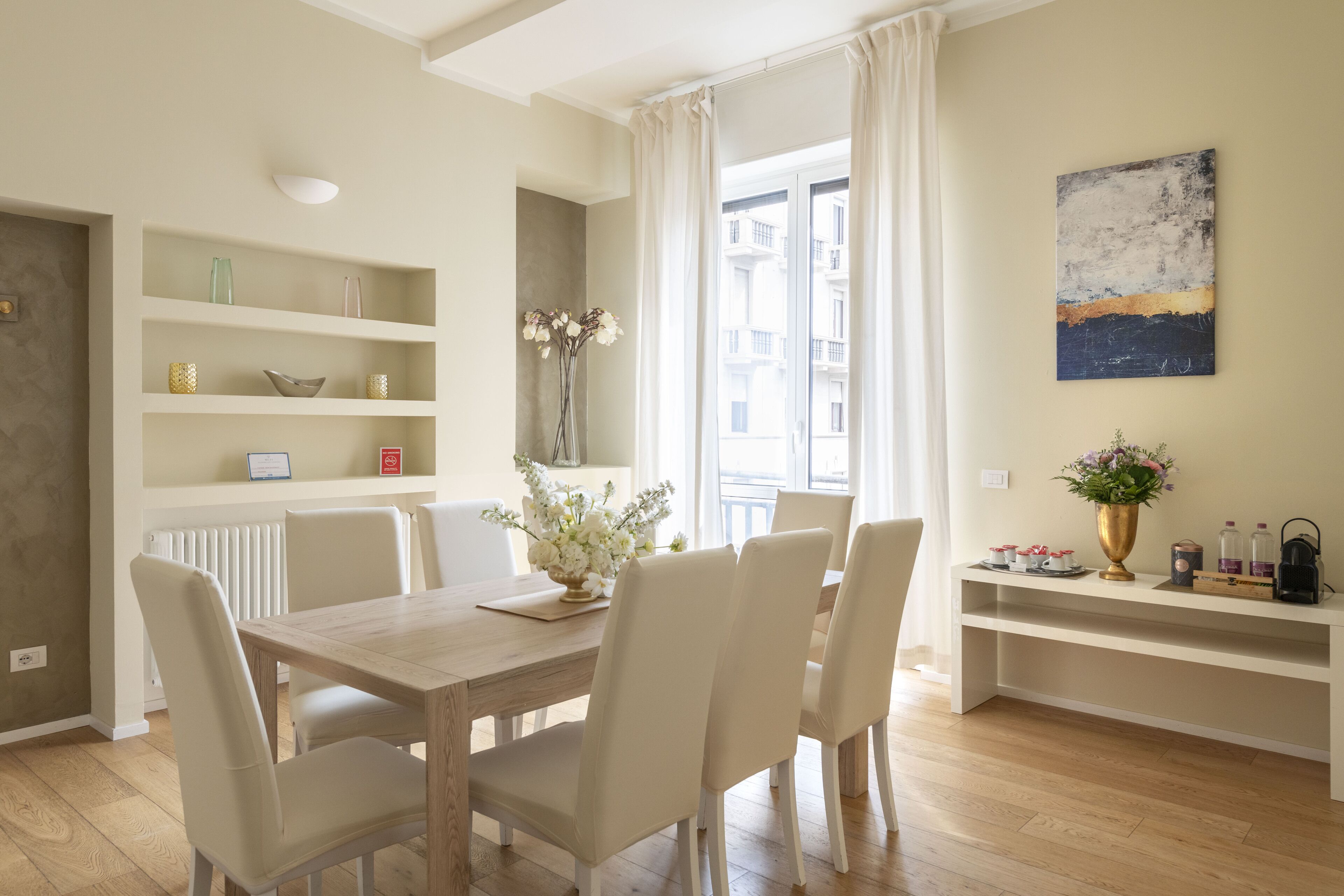 Un comedor con una mesa de madera y sillas blancas, una consola blanca con adornos, y vistas a edificios a través de una ventana con cortinas blancas.