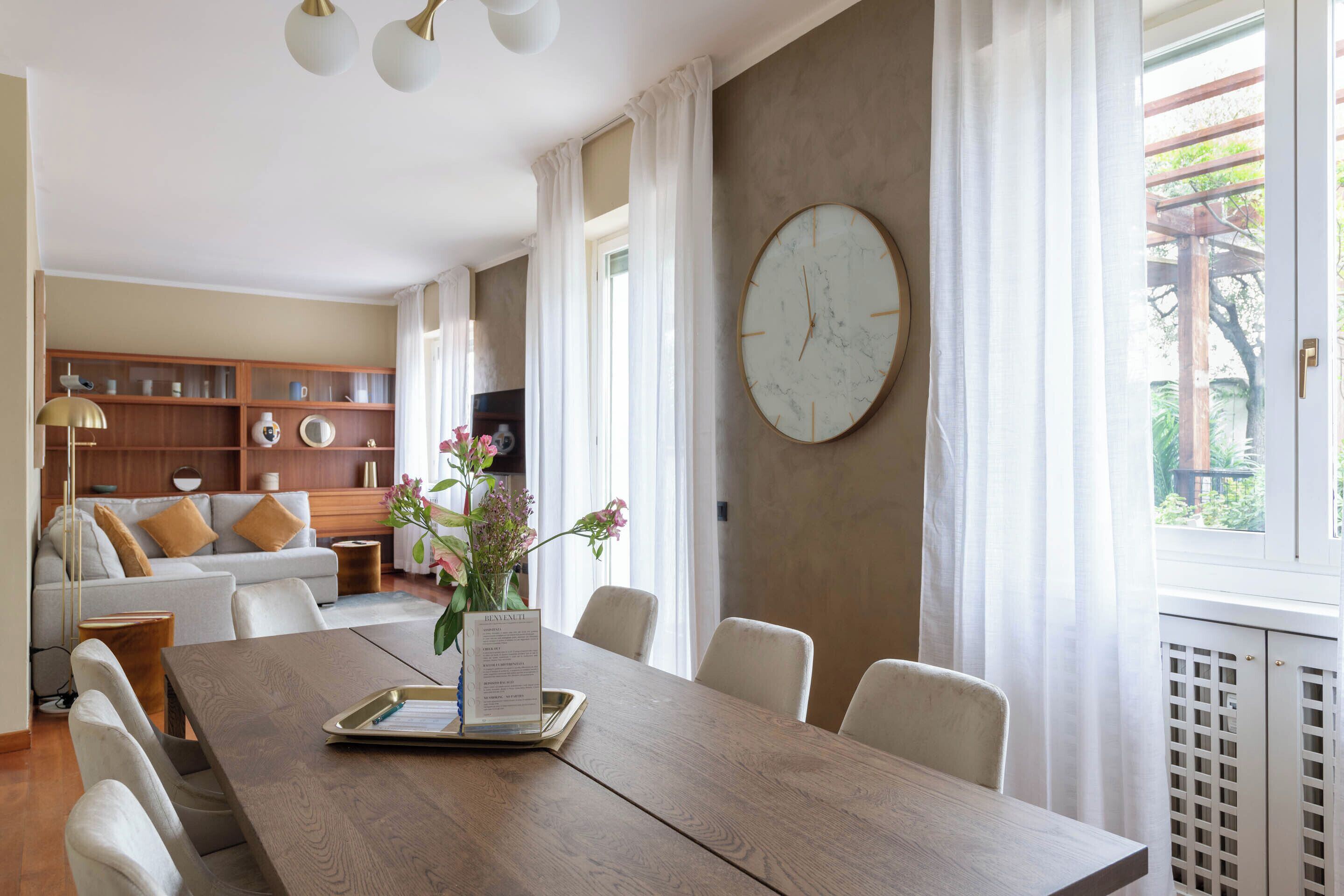 A dining area with a wooden table, white chairs, a large wall clock, and a vase with flowers.