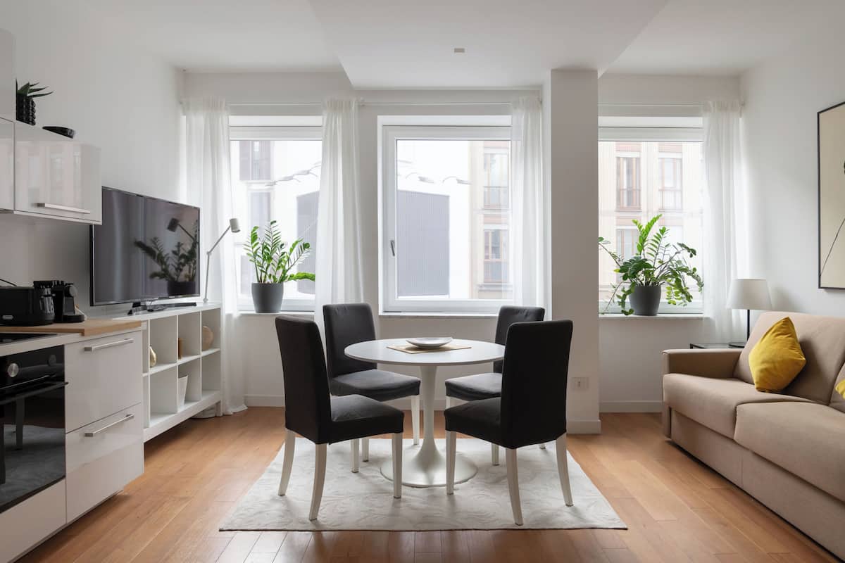Una sala de estar moderna con una mesa de comedor redonda, sillas negras, un sofá beige y un televisor sobre un mueble de madera.