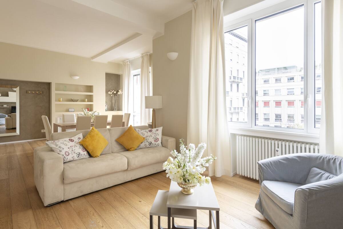 Un salón moderno con un sofá color beige, un sillón gris, una mesita con un jarrón de flores y un ventanal con vistas a la ciudad.