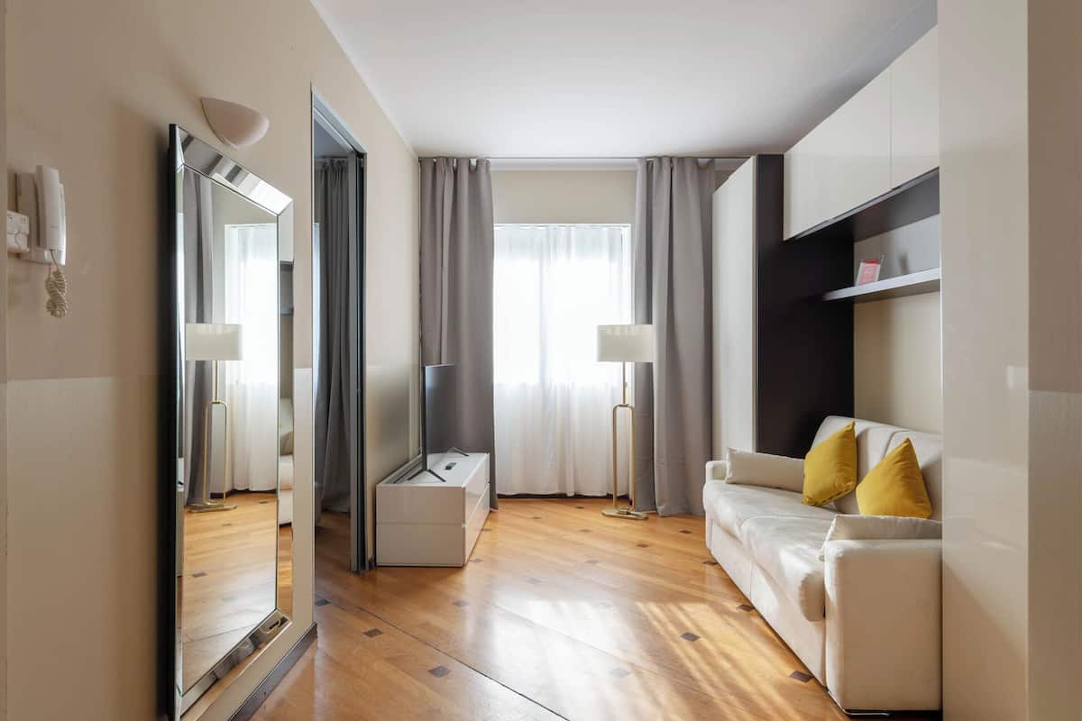 Una habitación de hotel moderna con piso de madera, un sofá blanco con cojines amarillos, una lámpara de pie, un espejo grande y un armario.