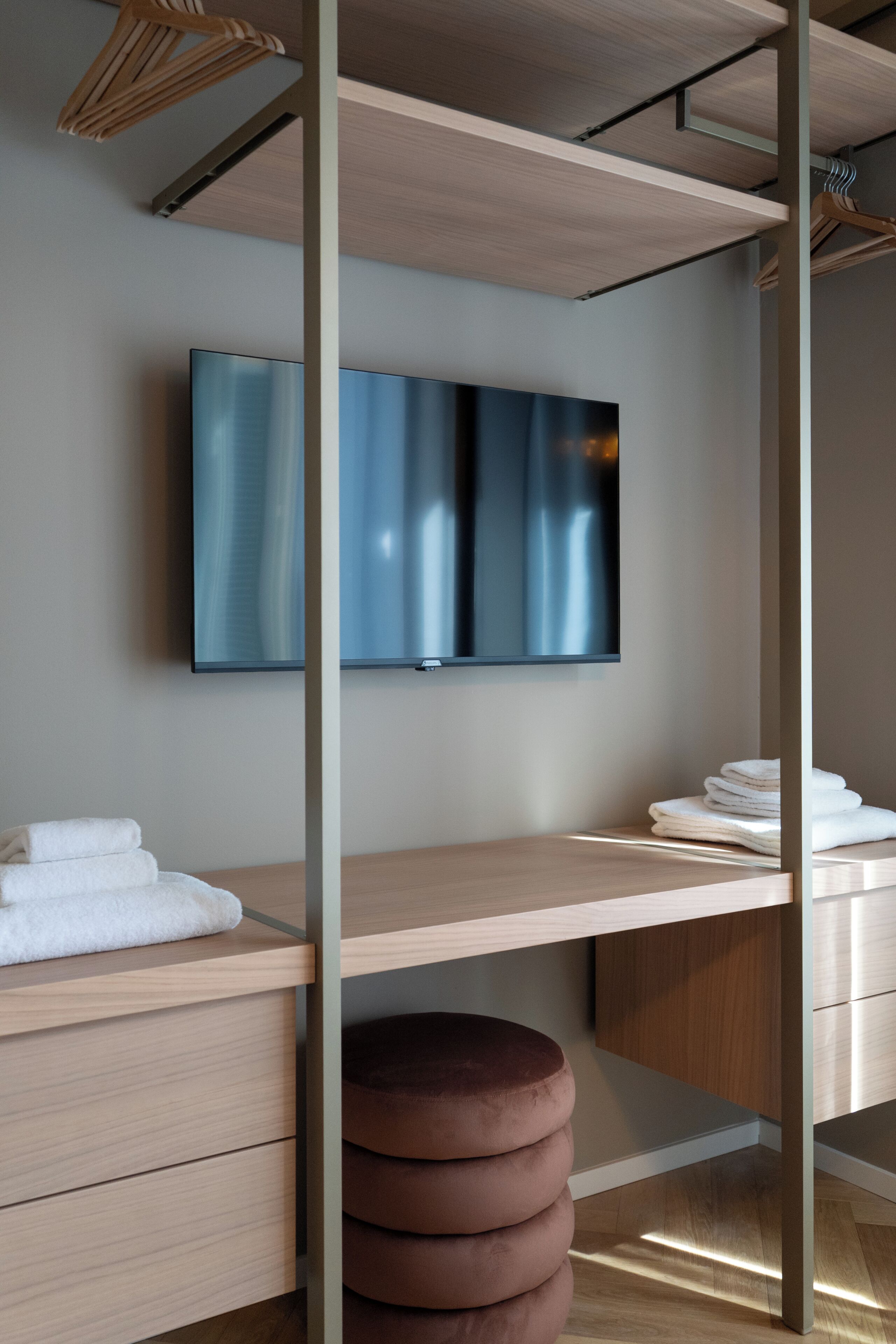 A modern room with a flat-screen TV mounted on the wall, a wooden desk with a cushioned stool, and a neatly folded towel on the desk.