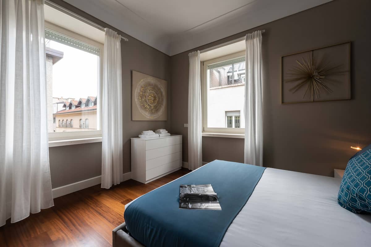 Un dormitorio con cama, cómoda y dos ventanas con cortinas.