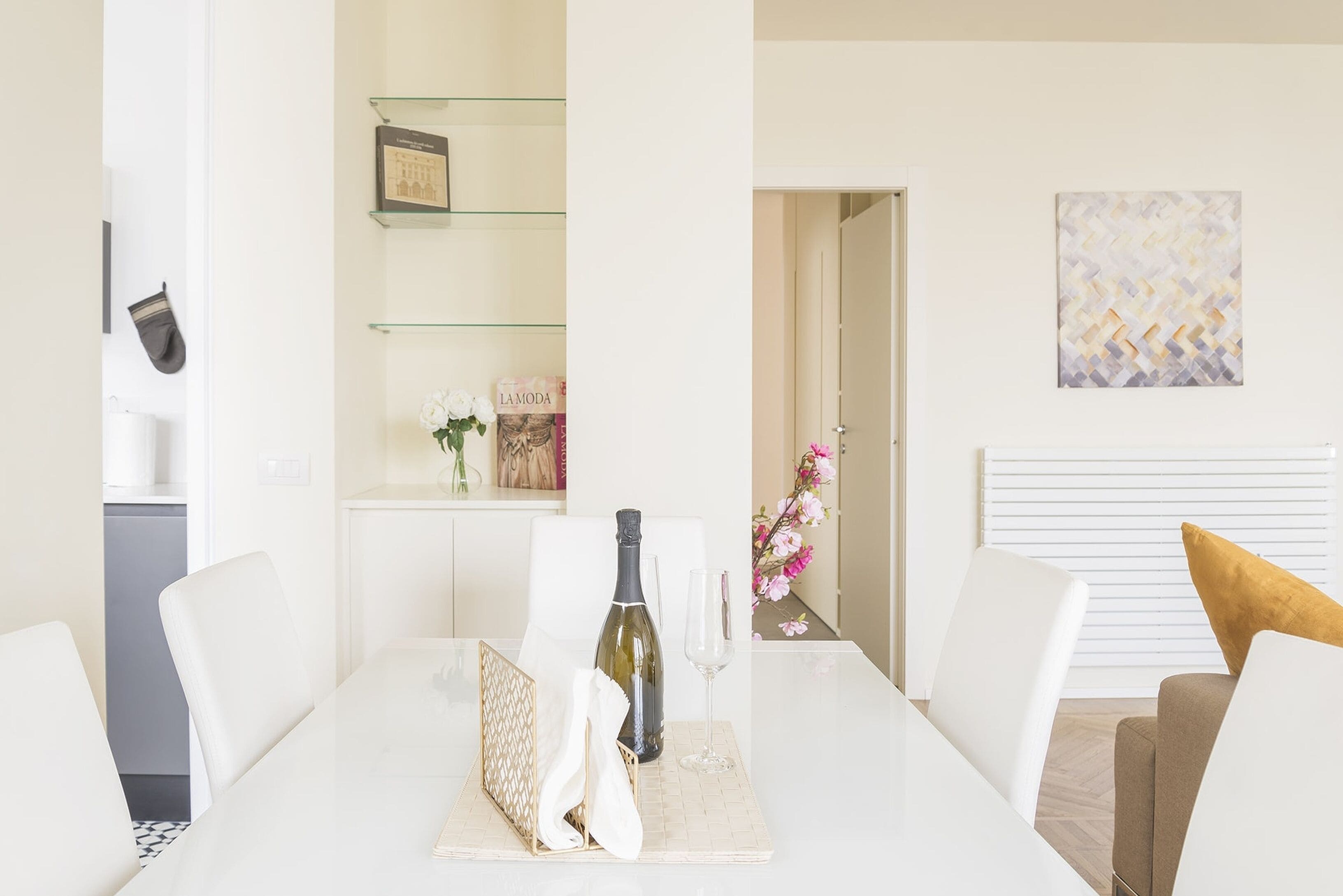 A modern dining area with a white table, chairs, a wine bottle, and a glass. There are shelves with decorative items and a framed picture on the wall.