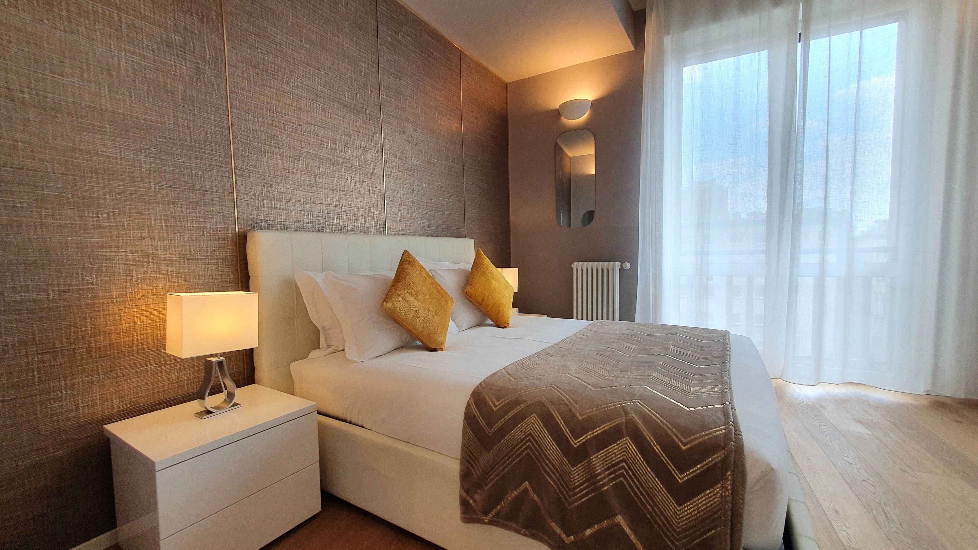 A hotel room with a large bed, a nightstand, a wall-mounted lamp, and a view of the outdoors through sheer curtains.