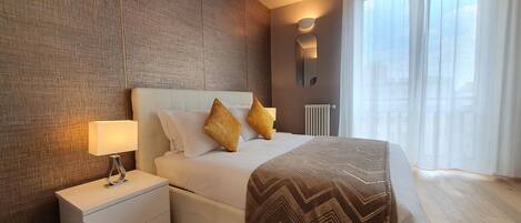 A hotel room with a large bed, a nightstand, a wall-mounted lamp, and a view of the outdoors through sheer curtains.