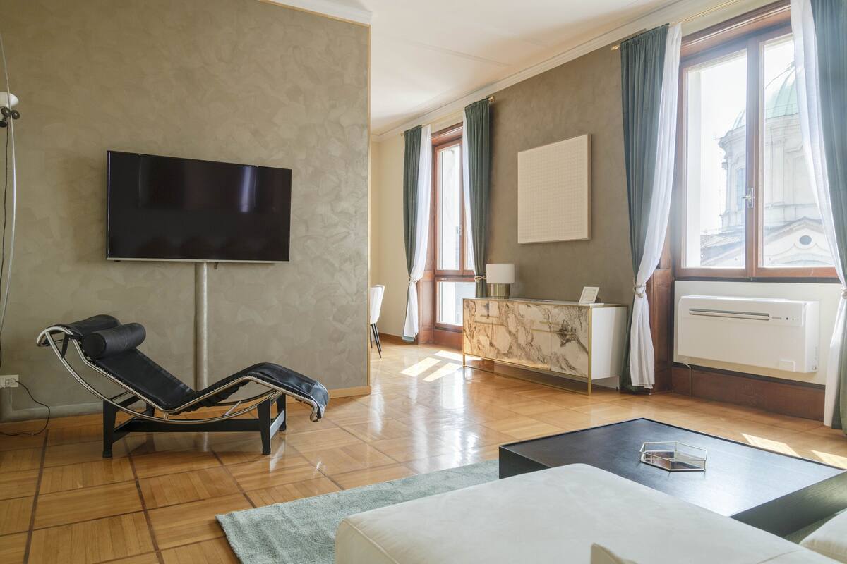 Una sala de estar moderna con un televisor de pantalla plana, un sillón de descanso, una recepción con mostrador de mármol y un amplio ventanal con cortinas.