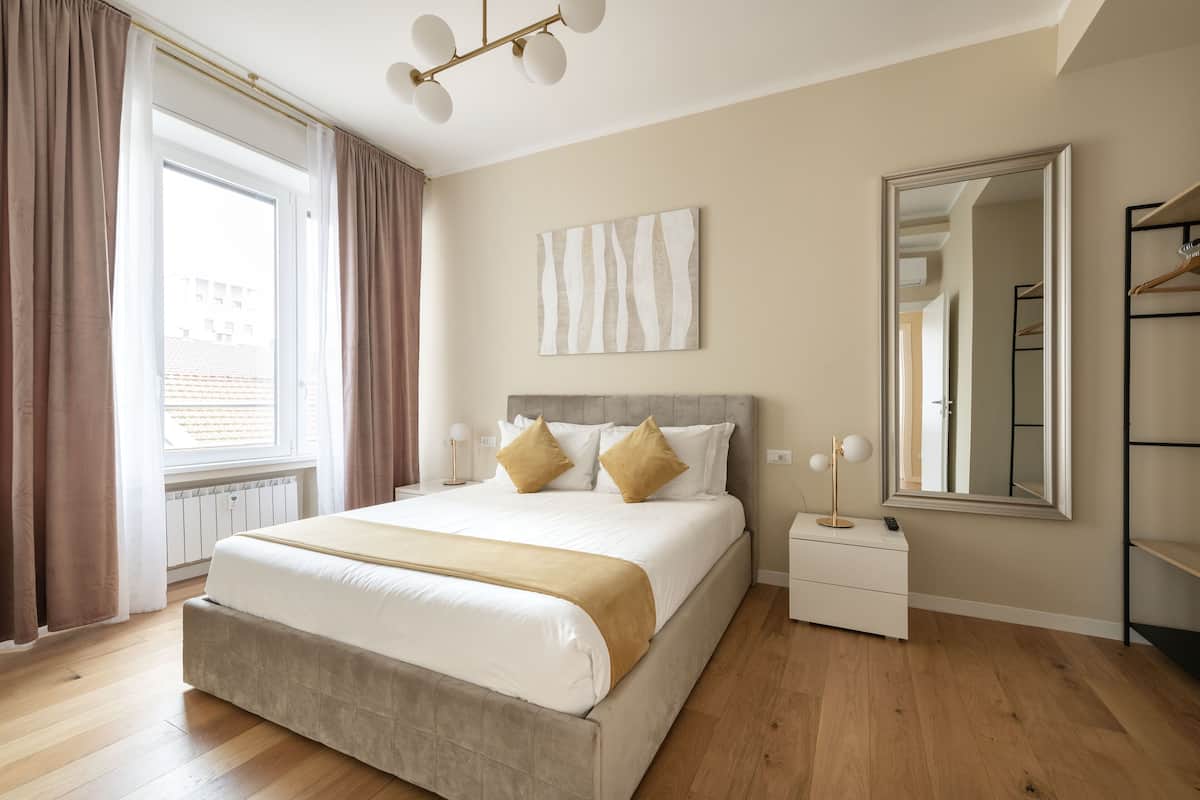 Un dormitorio con una cama grande, una mesita de noche, un espejo y una ventana con cortinas.