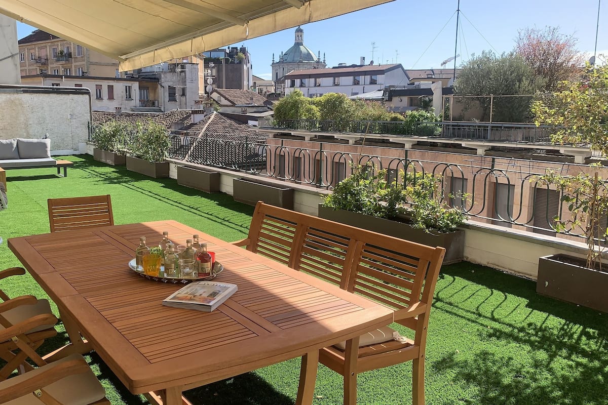 Una mesa de madera con sillas y una mecedora en una terraza en la azotea con césped artificial, con vistas a un paisaje urbano con edificios y una estructura en forma de cúpula.