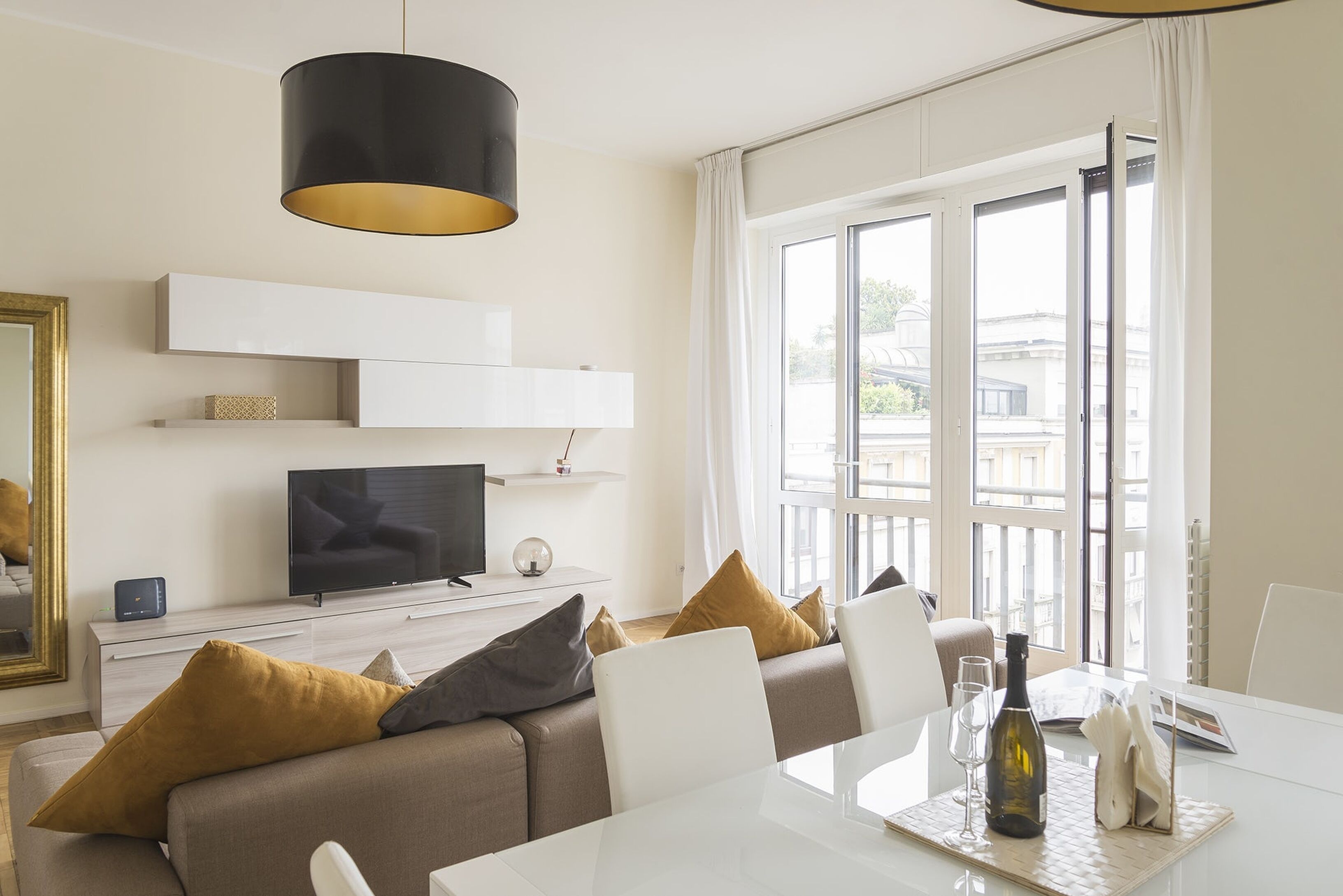Ein modernes Wohnzimmer mit einer Couch, einem Esstisch mit Flasche und Gläsern, einem Fernseher und einem großen Fenster mit Vorhängen.