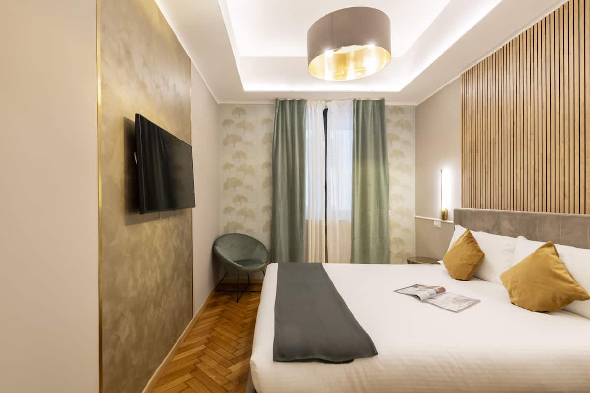 Habitación de hotel con cama, televisor de pantalla plana montado en la pared, una silla y una mesita de noche con lámpara.