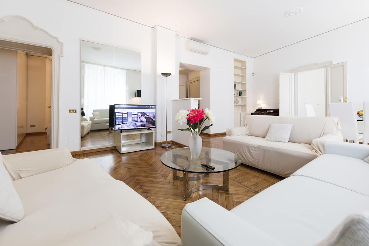 Una sala de estar moderna con una mesa de centro de vidrio, sofás blancos, un televisor y un jarrón con flores.