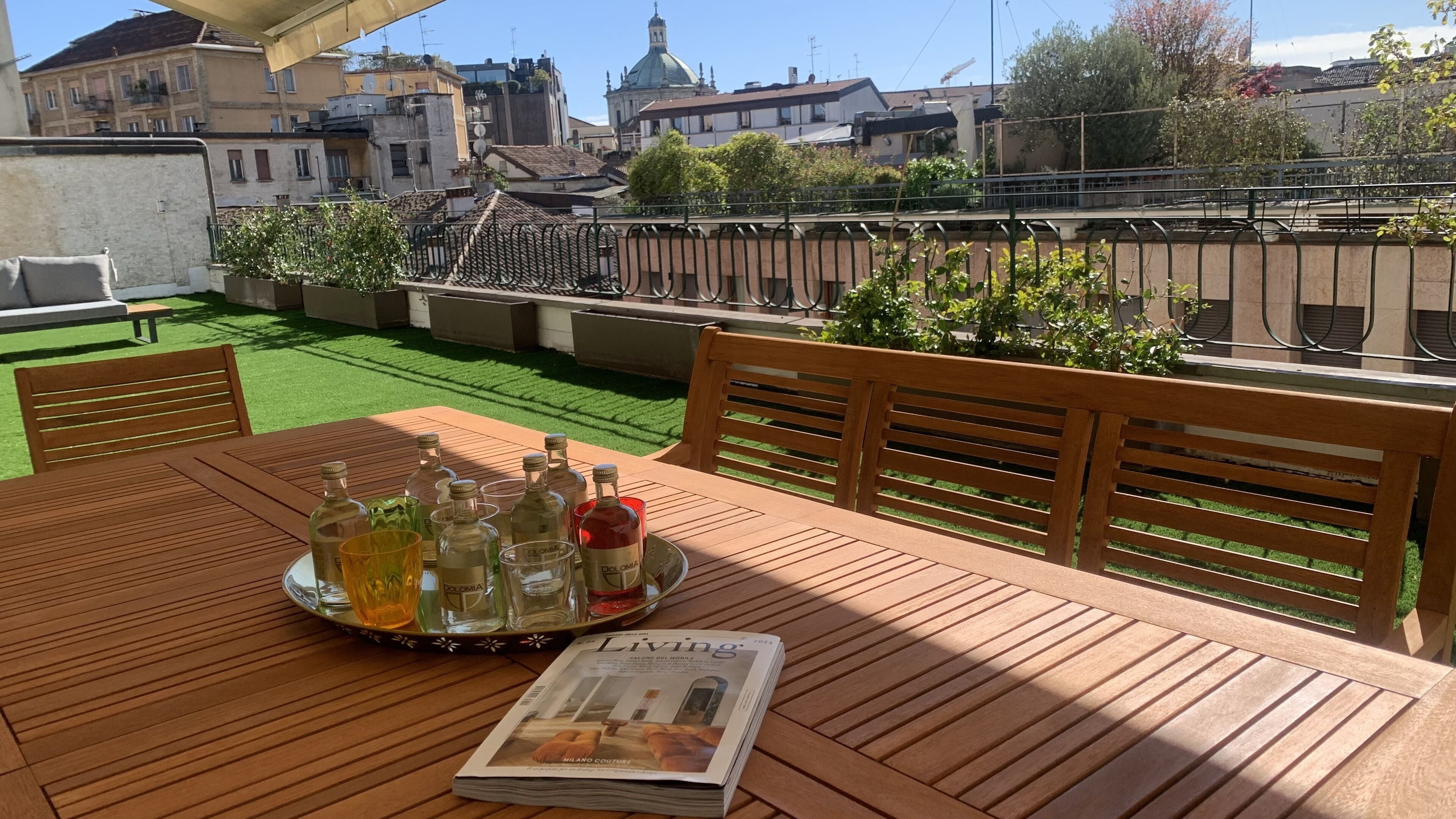 A wooden table with a tray of bottles and glasses, a newspaper, and a view of buildings and greenery.