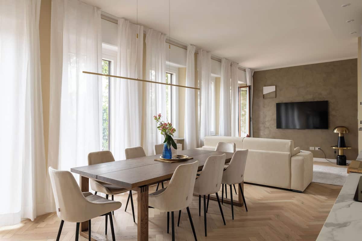 Una sala moderna con un sofá, una mesa de comedor con sillas, un televisor montado en la pared y un florero con flores sobre la mesa.