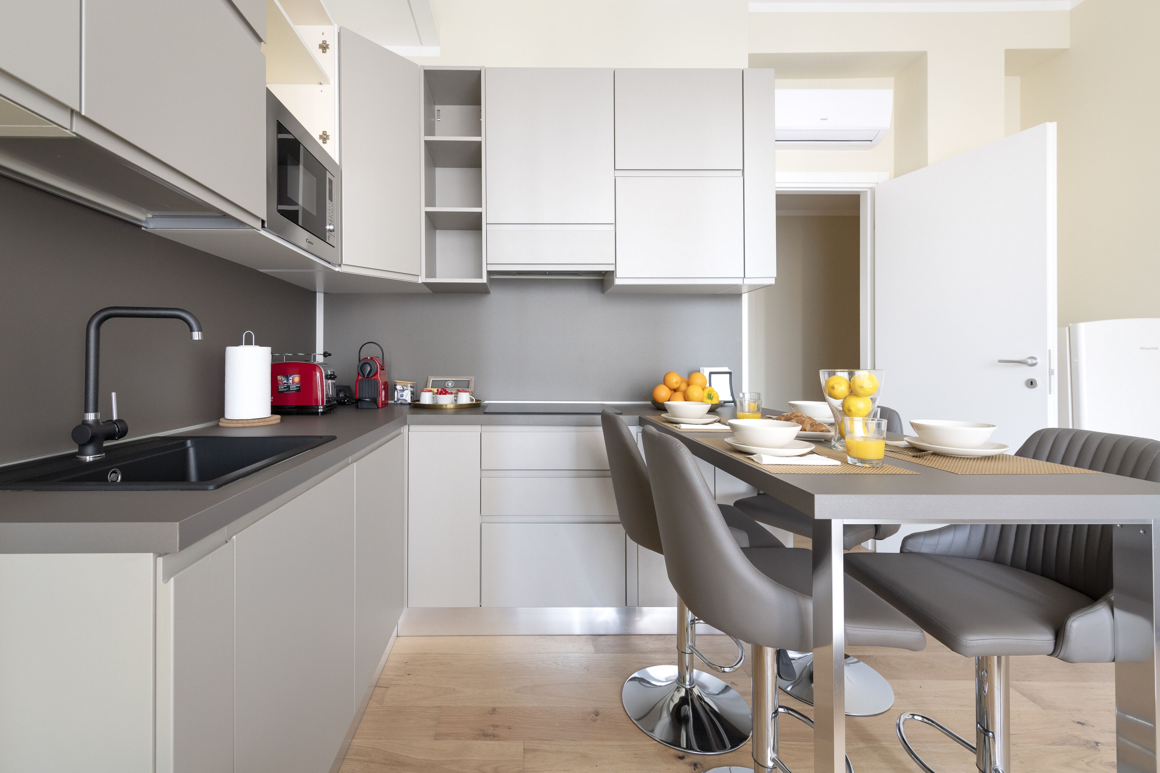 Una cocina moderna con gabinetes grises, un lavamanos negro y una zona de comedor con una mesa y taburetes de bar.
