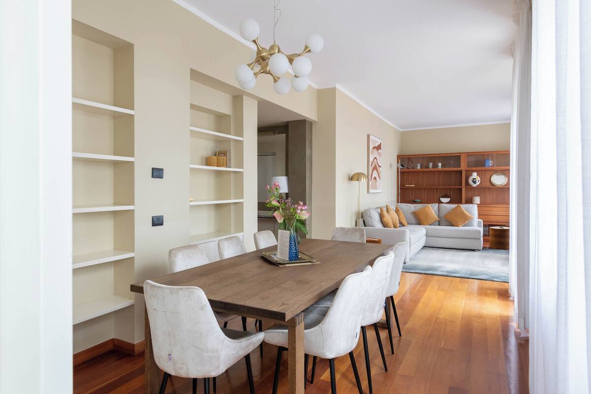 Un comedor con mesa de madera y sillas blancas, una estantería, un sofá y un jarrón decorativo con flores.