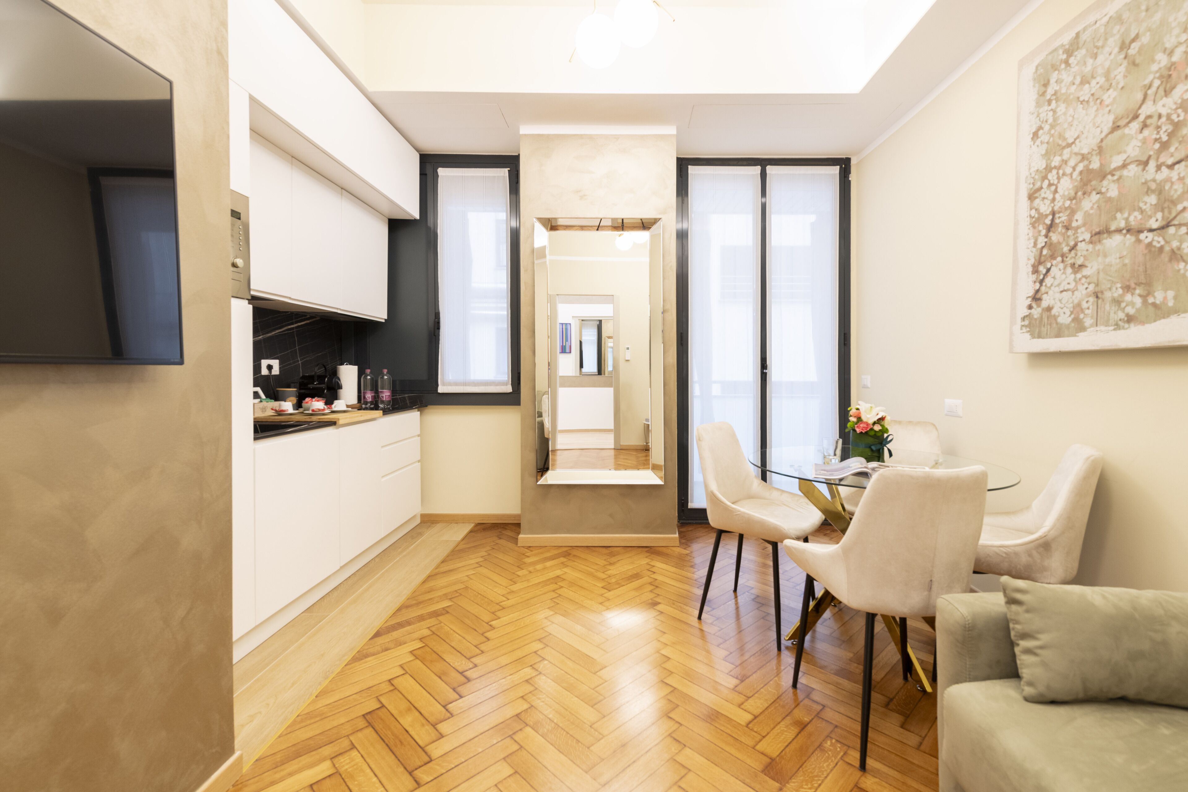 A modern kitchen with white cabinets, a wooden herringbone floor, and a dining area with a glass table and white chairs.