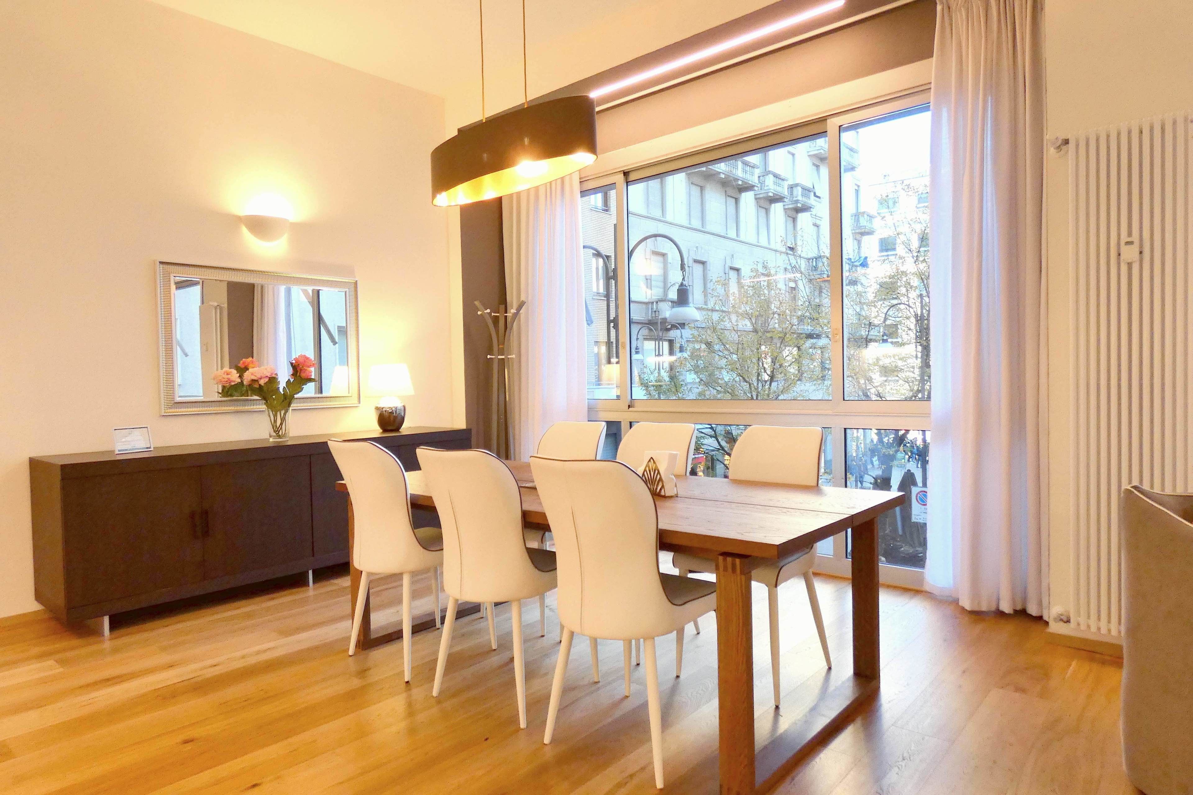 Un comedor moderno con una mesa de madera y sillas blancas, una consola oscura, un ventanal con vistas a edificios y una lámpara colgante.
