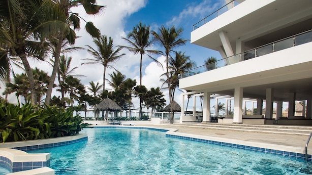 A swimming pool with clear blue water, surrounded by palm trees and a modern building with balconies.