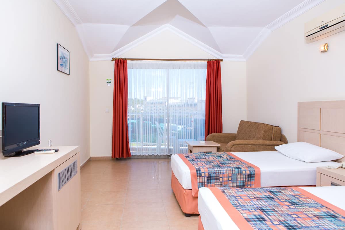 A hotel room with two beds, a TV, a desk, and a balcony with red curtains.