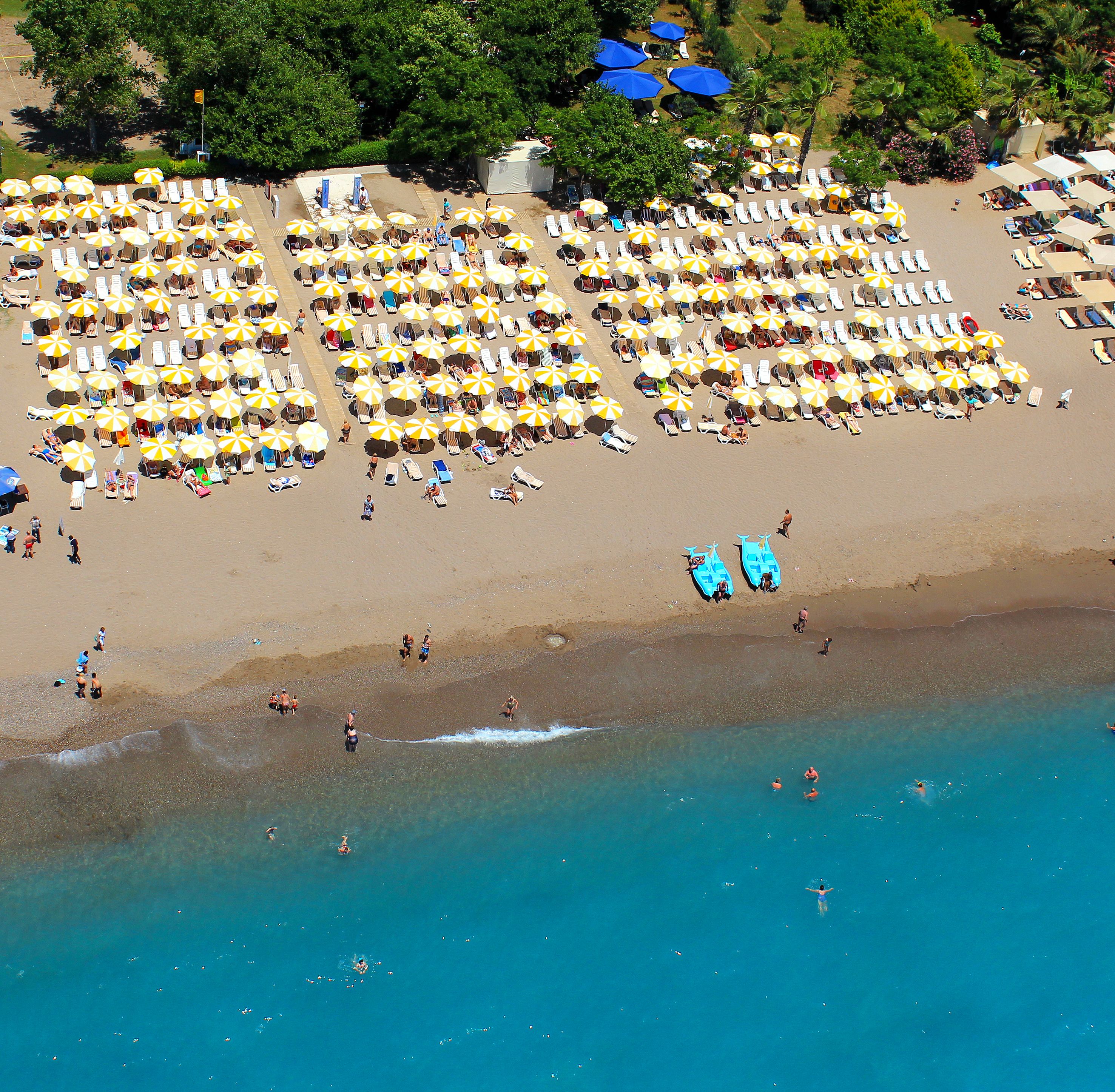 An aerial view of a beach with numerous sun loungers and umbrellas, a few people swimming in the water, and a few boats near the shore.