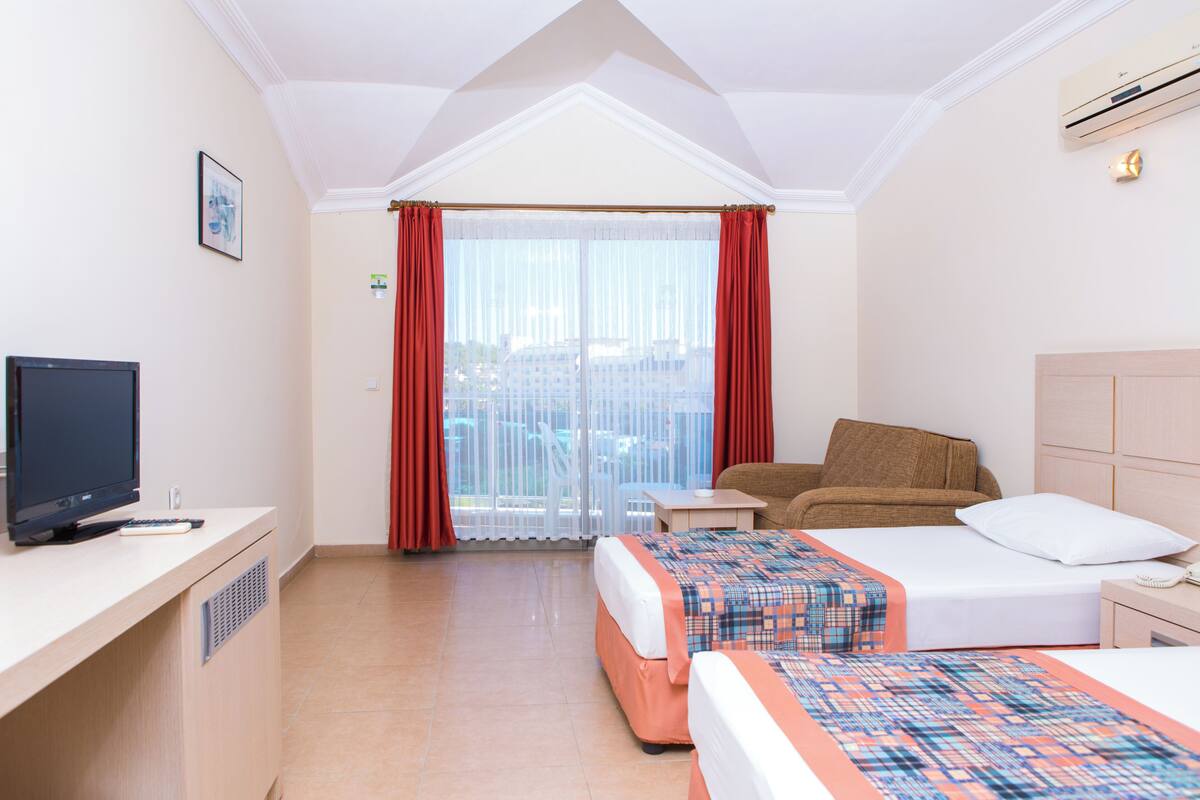 A hotel room with two beds, a TV, a desk, and a balcony with red curtains.