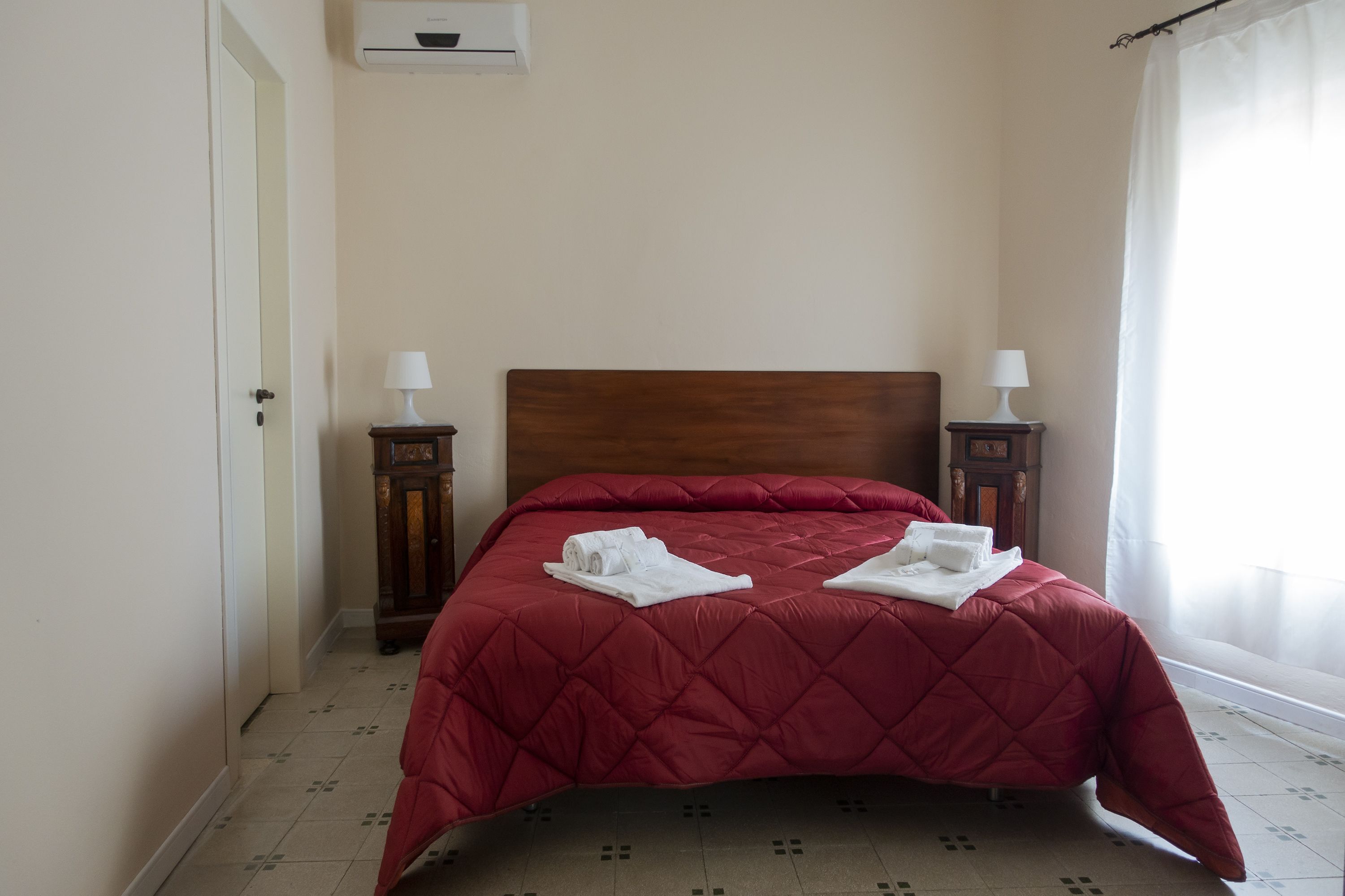 Una cama bien hecha con colcha roja, dos mesitas de noche con lámparas y una ventana con cortinas.