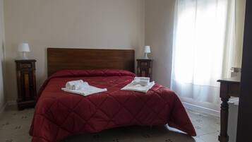 Una cama bien hecha con colcha roja, dos mesitas de noche con lámparas y una ventana con cortinas traslúcidas.