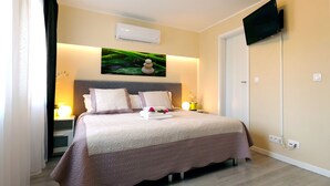 A neatly made bed with a patterned bedspread, a wall-mounted air conditioner, a flat-screen TV, and a bedside table with a lamp and a vase of flowers. - My-Cocoon (Court-Saint-Etienne)