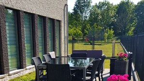 A patio with a glass table and chairs, surrounded by a wooden deck and a brick planter with flowers. - My-Cocoon (Court-Saint-Etienne)