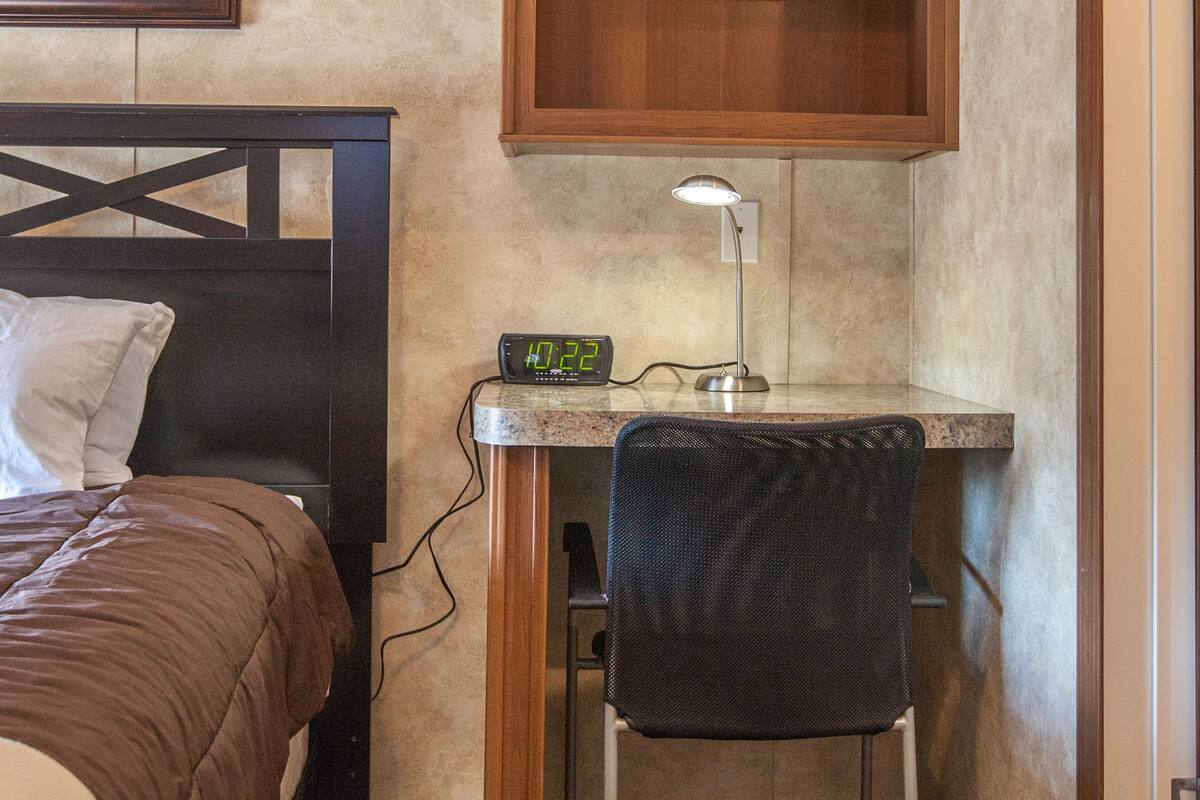 A bedroom with a bed, a desk with a chair, a clock, and a shelf above the desk.