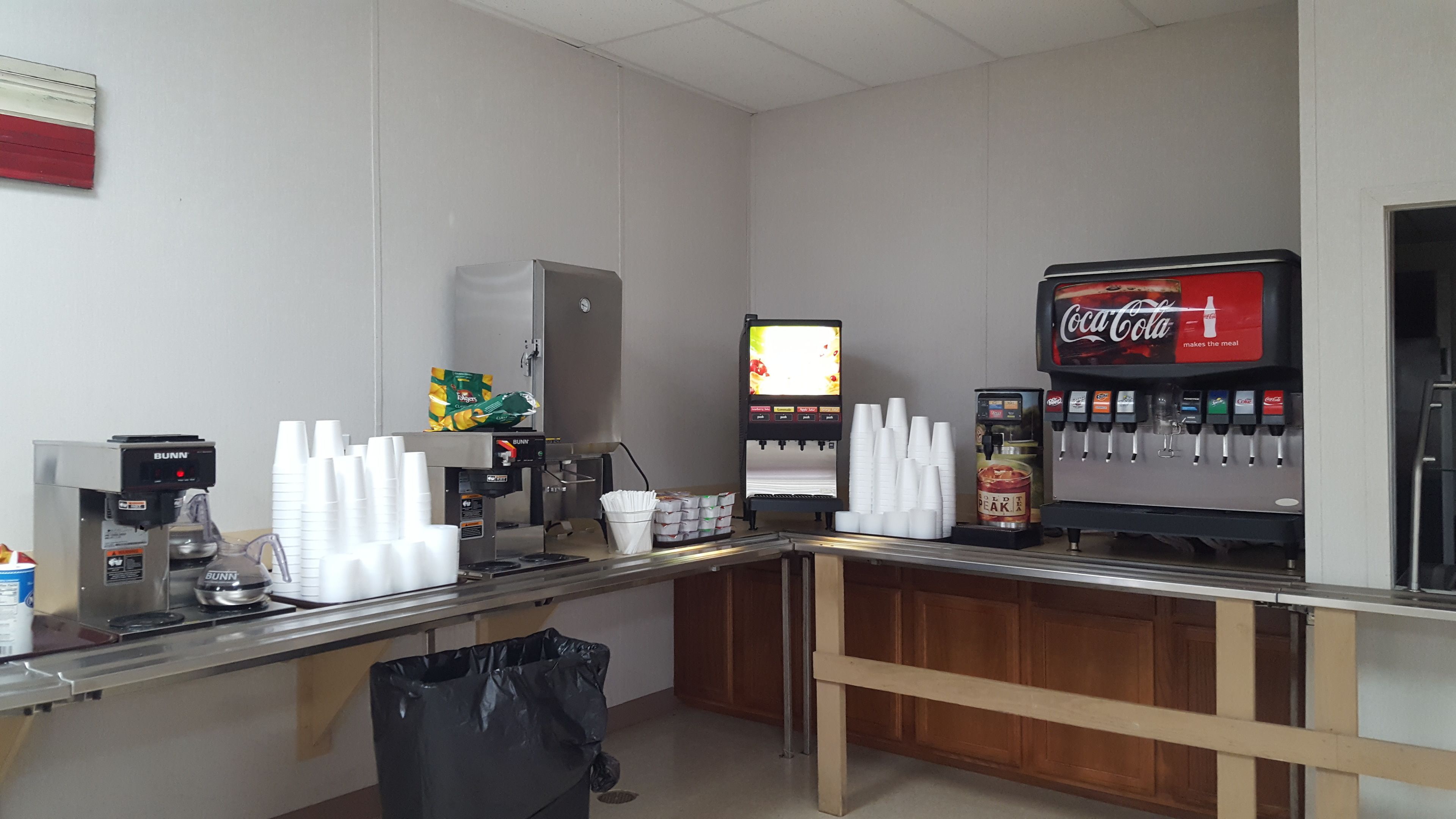 A self-service beverage station with multiple drink dispensers, including Coca-Cola and other sodas, and a coffee machine.